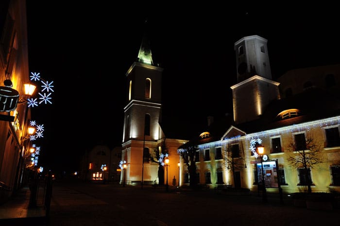 Nocny rynek z podświetloną wieżą kościoła i pierzeją kamienic; na latarni z lewej białe dekoracje w kształcie śnieżynek